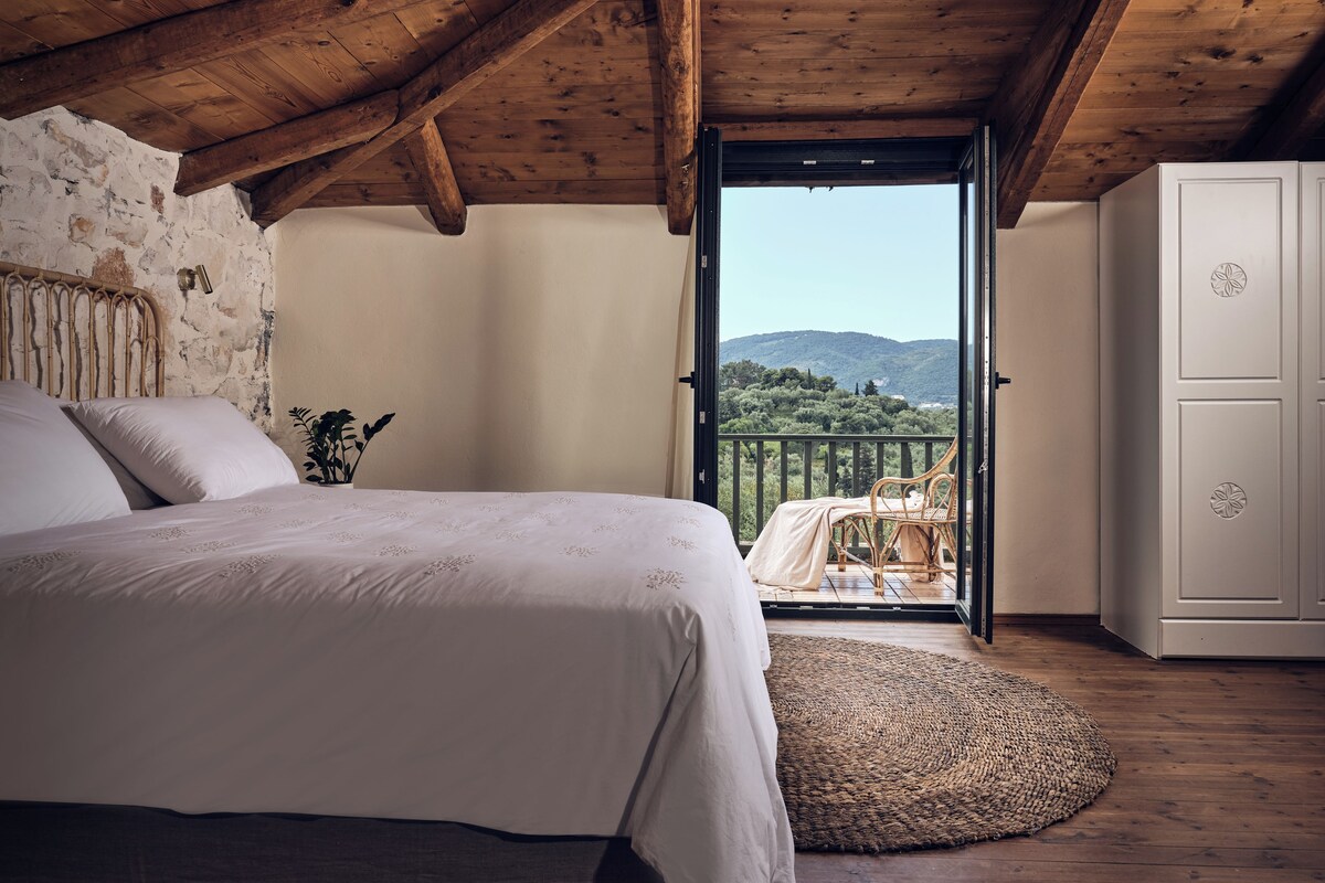 A bedroom with a wooden ceiling, a bed with white linens, a view of greenery and hills through an open door, and a wicker chair outside.