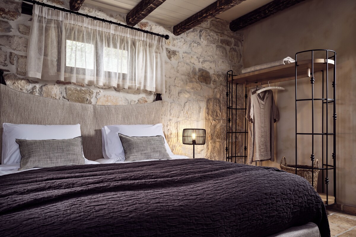 A bedroom with a stone wall, a wooden ceiling, a bed with a dark blanket, a bedside lamp, and a metal rack with clothes.