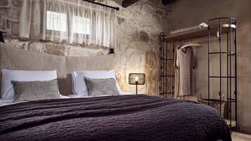 A bedroom with a stone wall, a wooden ceiling, a bed with a dark blanket, a bedside lamp, and a metal rack with clothes.