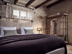 A bedroom with a stone wall, a wooden ceiling, a bed with a dark blanket, a bedside lamp, and a metal rack with clothes.