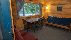Un accogliente interno di una baita con un tavolo da pranzo in legno, quattro sedie, un divano blu e una finestra con tende.