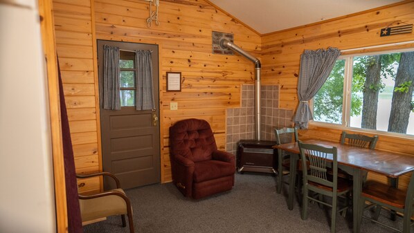 L’intérieur d’un chalet accueillant, avec une porte en bois, un fauteuil moelleux, une table à manger avec des chaises et une fenêtre avec des rideaux.