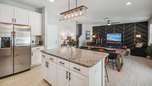 A modern kitchen with a central island, stainless steel refrigerator, and a dining area with a TV and seating.