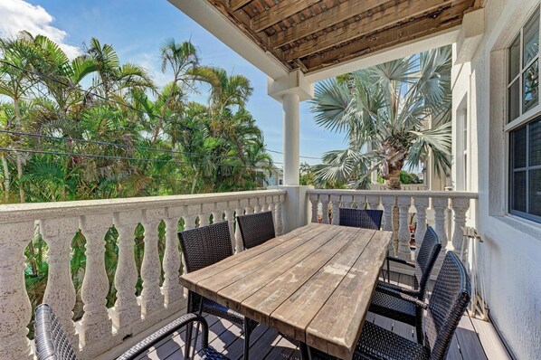 A wooden table with chairs on a balcony with a white balustrade, overlooking palm trees and a clear sky.