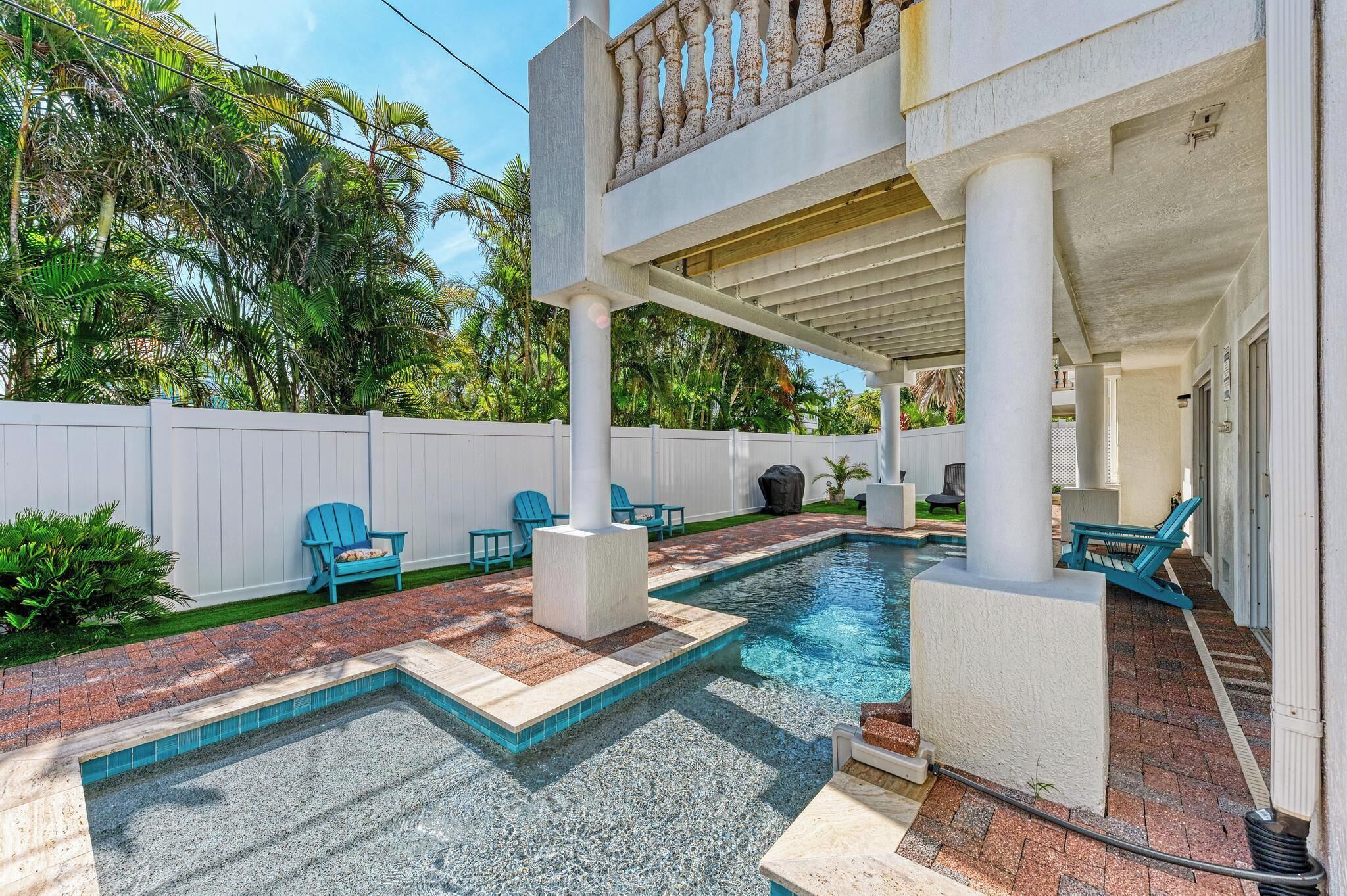 A swimming pool with a brick deck and blue chairs, surrounded by a white fence and palm trees.