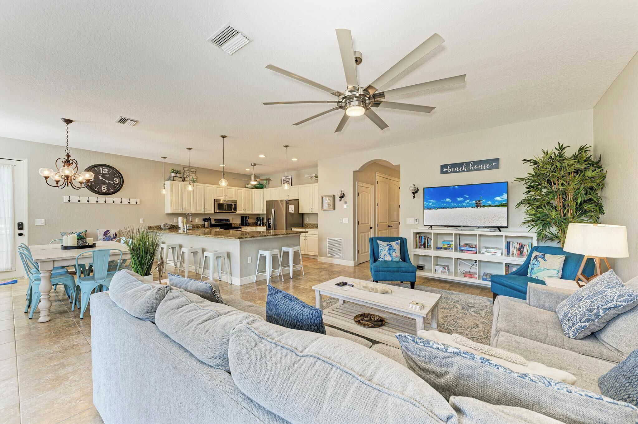 A modern living room with a large gray sofa, a coffee table, and a blue armchair. There is a kitchen area with white cabinets and a dining table with blue chairs. A ceiling fan and a TV are visible in the background.