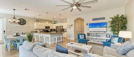 A modern living room with a large gray sofa, a coffee table, and a blue armchair. There is a kitchen area with white cabinets and a dining table with blue chairs. A ceiling fan and a TV are visible in the background.