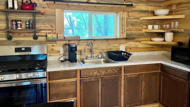 Cocina compacta con armarios de madera, estufa de acero inoxidable, un lavamanos y una ventana con vistas a árboles.
