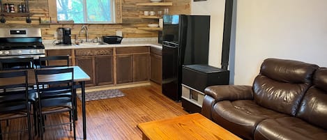 A cozy kitchen with wooden cabinets, a stove, and a black refrigerator. There is a dining table with chairs and a leather sofa.