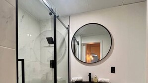 Moderni kylpyhuone, jossa on lasinen suihkuseinä, puinen allaskaappi pyöreällä peilillä sekä valkoinen wc-istuin.