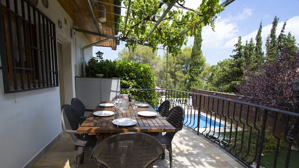 En udendørs terrasse med et træbord dækket til spisning, omgivet af stole, og en pool i baggrunden.