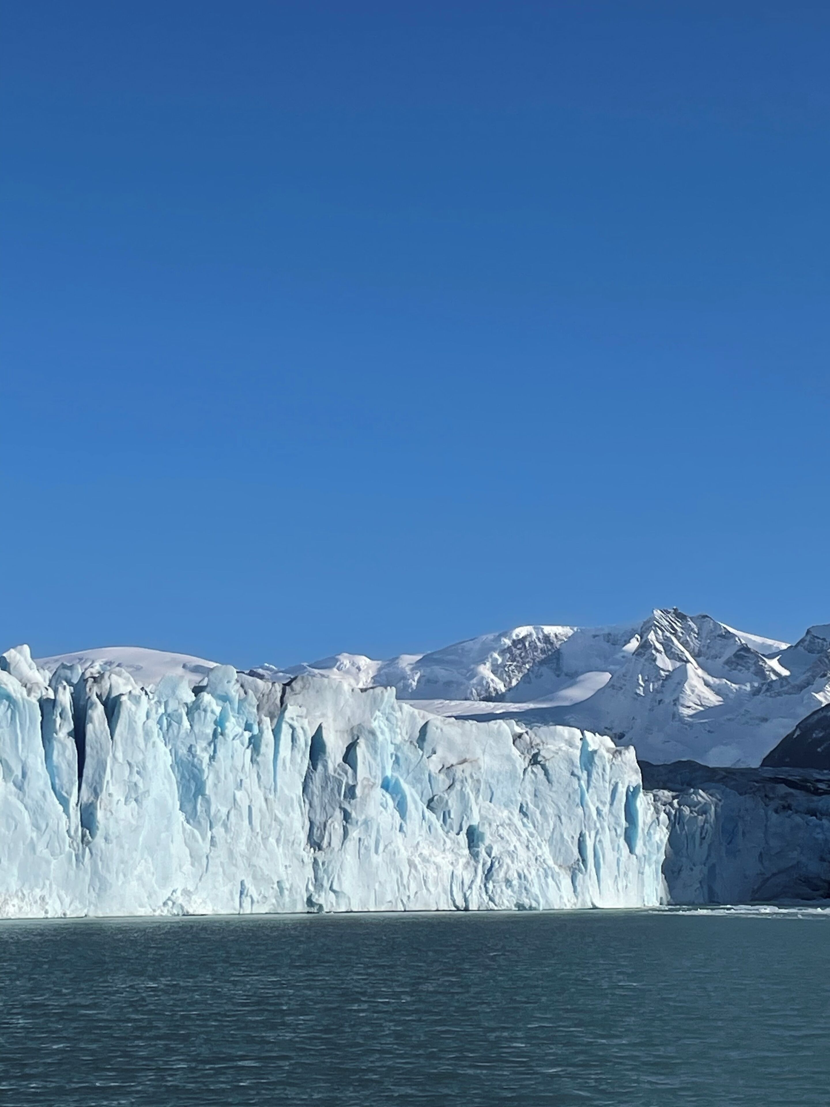 A glacier with a clear view of its massive ice formations and a distant mountain range under a clear blue sky.