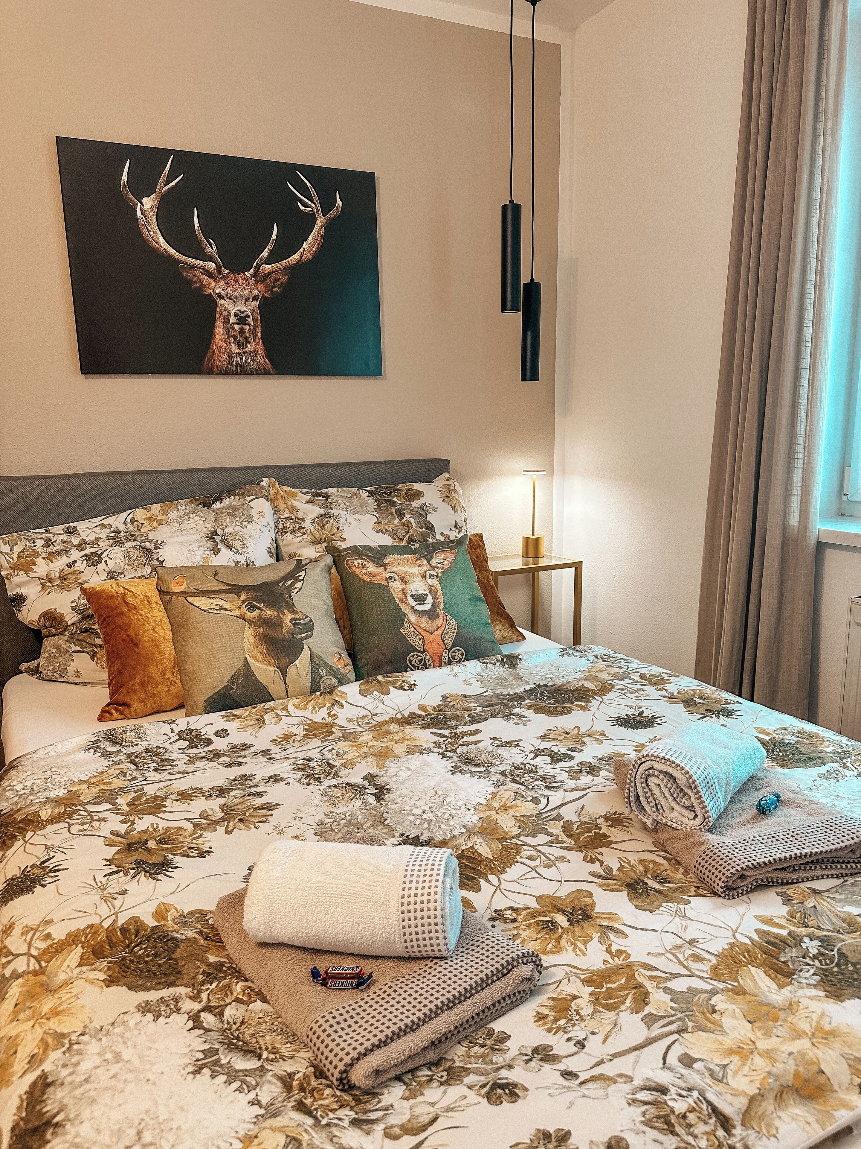 A neatly made bed with floral bedding, a deer-themed pillow, and a deer antler painting on the wall.