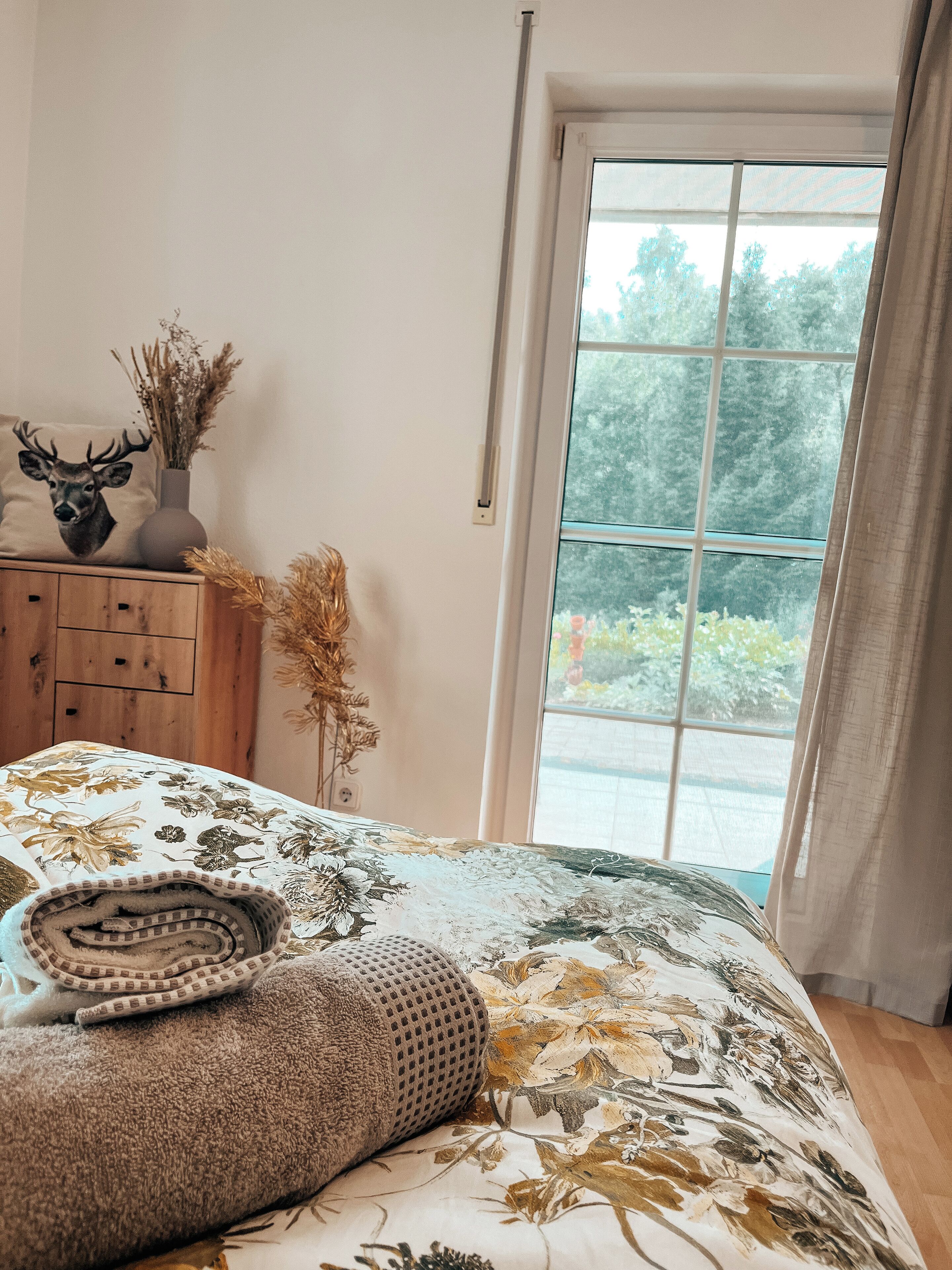 Un dormitorio con colcha estampada, cómoda de madera y cojín con forma de ciervo, y una ventana con vista a árboles.
