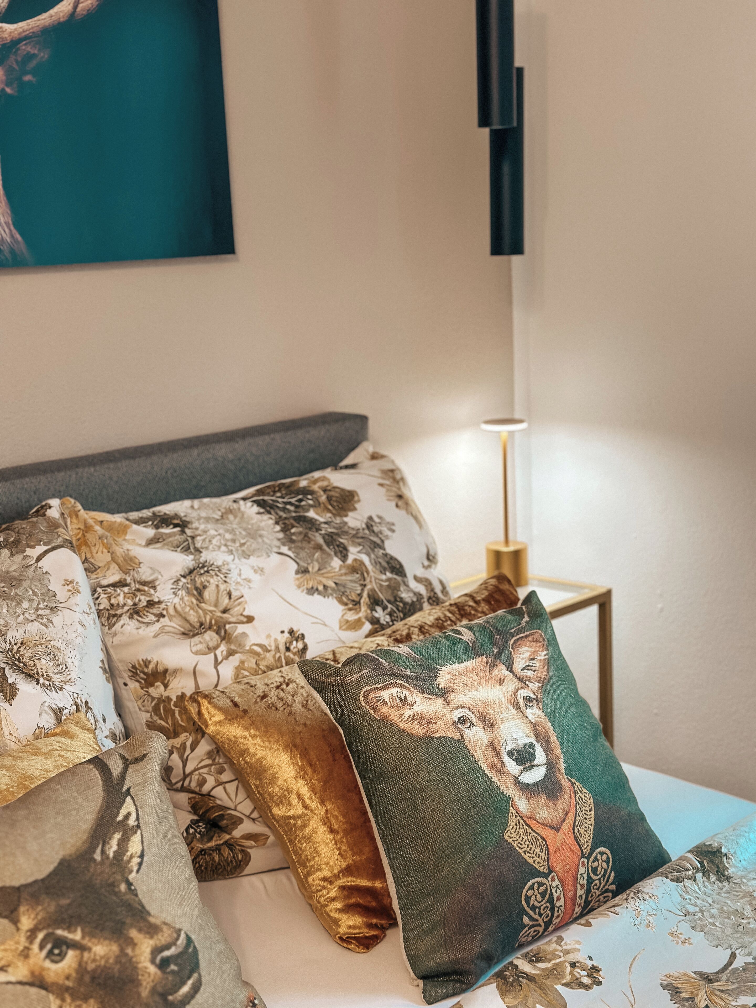 Un lit bien fait, agrémenté d’oreillers aux motifs floraux et animaliers, une lampe de chevet moderne et une œuvre d’art encadrée au mur.