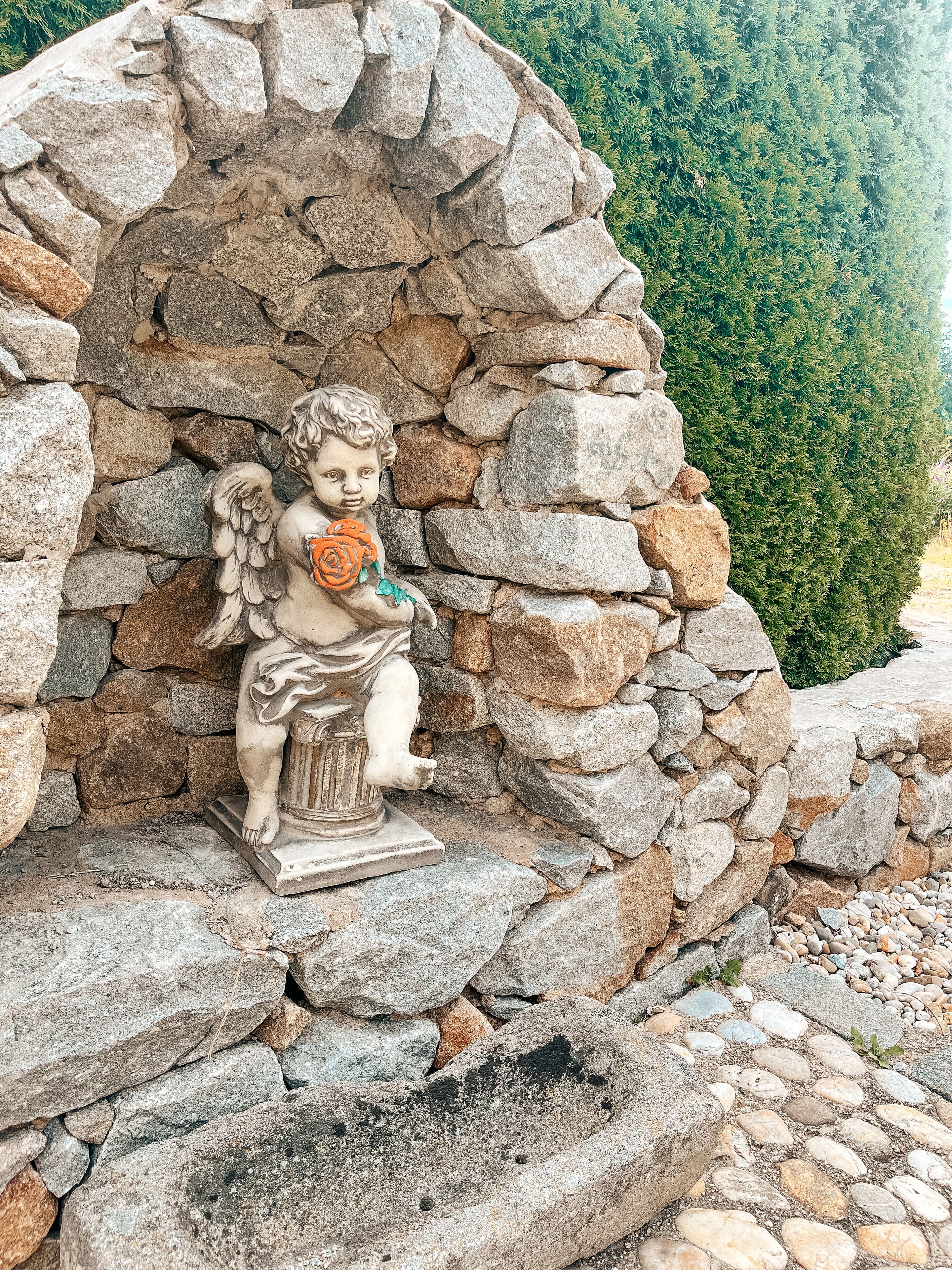 Une statue en pierre représentant un enfant tenant une fleur, placée dans une alcôve rocheuse, avec une haie verte en arrière-plan.