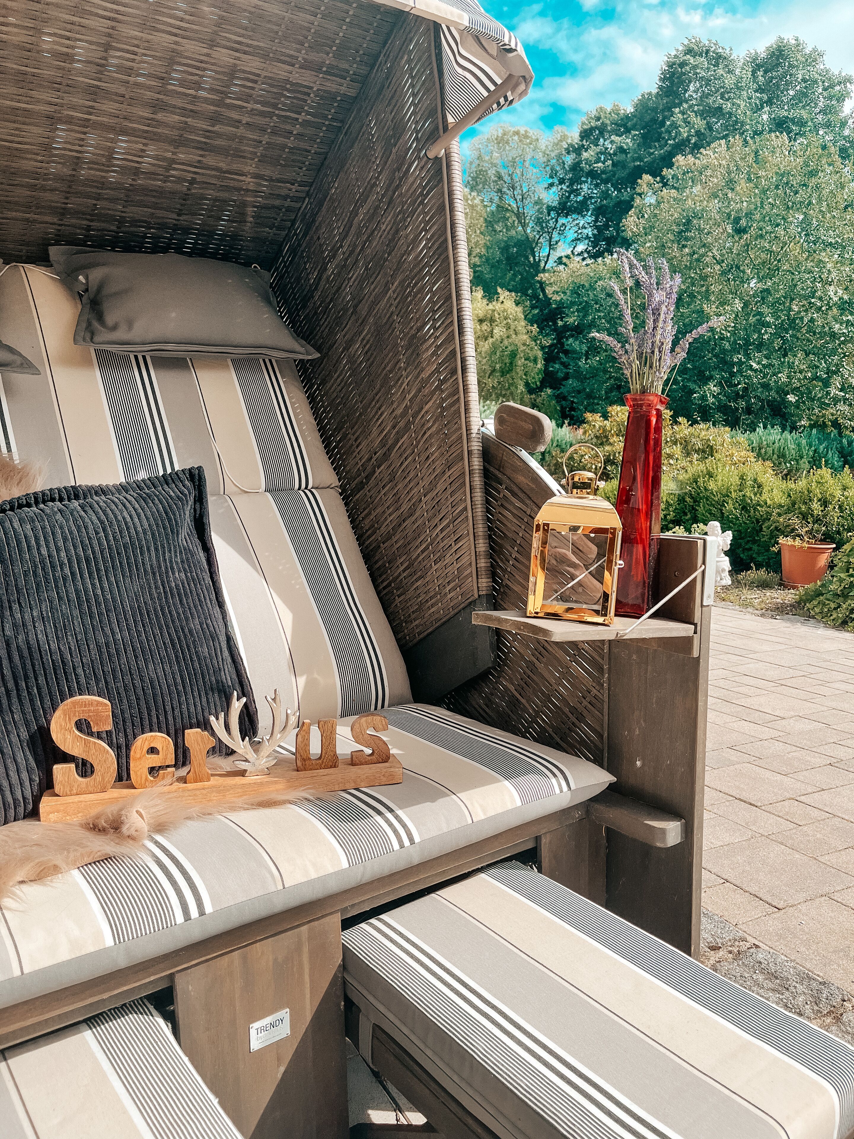 An outdoor seating area with a cushioned bench, a wooden sign reading "Serus," and a vase with lavender.