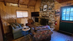 A cozy cabin interior with a stone fireplace, a sofa, a patterned armchair, a lamp, and a television. - Rustic Lakeview Lodge at Bass Lake. Bookable boat slip! (Bass Lake)