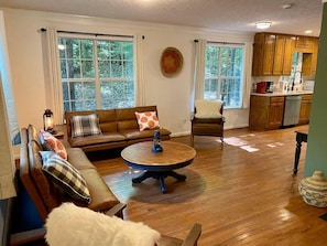 A spacious living room with a wooden floor, a round coffee table, a brown leather sofa, and a chair. There is a kitchen area with wooden cabinets and a dining table. - Gold Dust Getaway - Close to town, Fire Pit (Dahlonega)