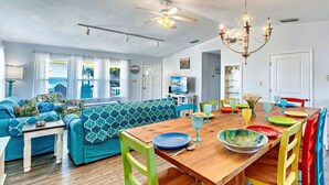 A bright living room with a wooden dining table, colorful chairs, and a blue patterned sofa. - Beach Shack - Across Street From Beach, Private Swimming Pool, Pet Friendly! (St. George Island)