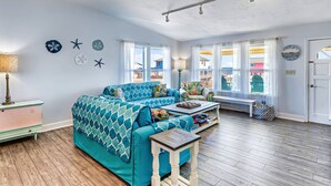 A cozy living room with a teal sofa, wooden flooring, and a view of the beach through large windows. - Beach Shack - Across Street From Beach, Private Swimming Pool, Pet Friendly! (St. George Island)