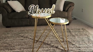 Un salon avec un canapé marron foncé, une table basse ronde en verre aux pieds dorés et un panneau décoratif sur lequel on peut lire le mot « Béni ».