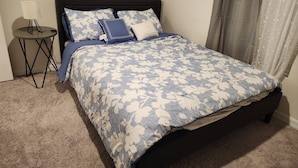 Un lit bien fait, recouvert d’une couvre-lit à motifs floraux, une petite table de chevet avec une lampe et une fenêtre avec des rideaux.