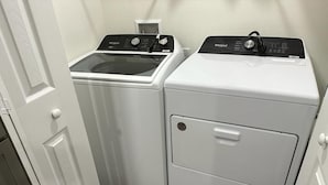Une petite buanderie équipée d’un lave-linge et d’un sèche-linge à chargement frontal, d’une petite étagère au-dessus du sèche-linge et d’une porte donnant sur une autre pièce.