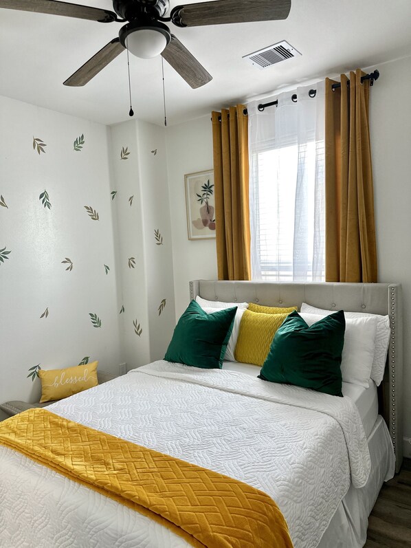 Una cama bien hecha con colcha blanca y amarilla, cabecera tapizada de gris y ventilador de techo.
