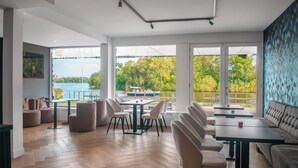 A modern dining area with a view of a lake and boats, featuring wooden flooring, contemporary furniture, and large windows. - Amsterdam Lake Hotel  (Badhoevedorp)