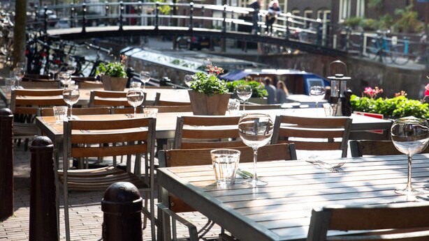 An outdoor dining area with wooden tables and chairs, potted plants, and a canal view. - Amsterdam Lake Hotel  (Badhoevedorp)