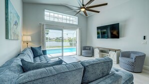 Una sala moderna con un sofá seccional gris grande, un ventilador de techo, un televisor y vistas a una piscina a través de amplios ventanales.