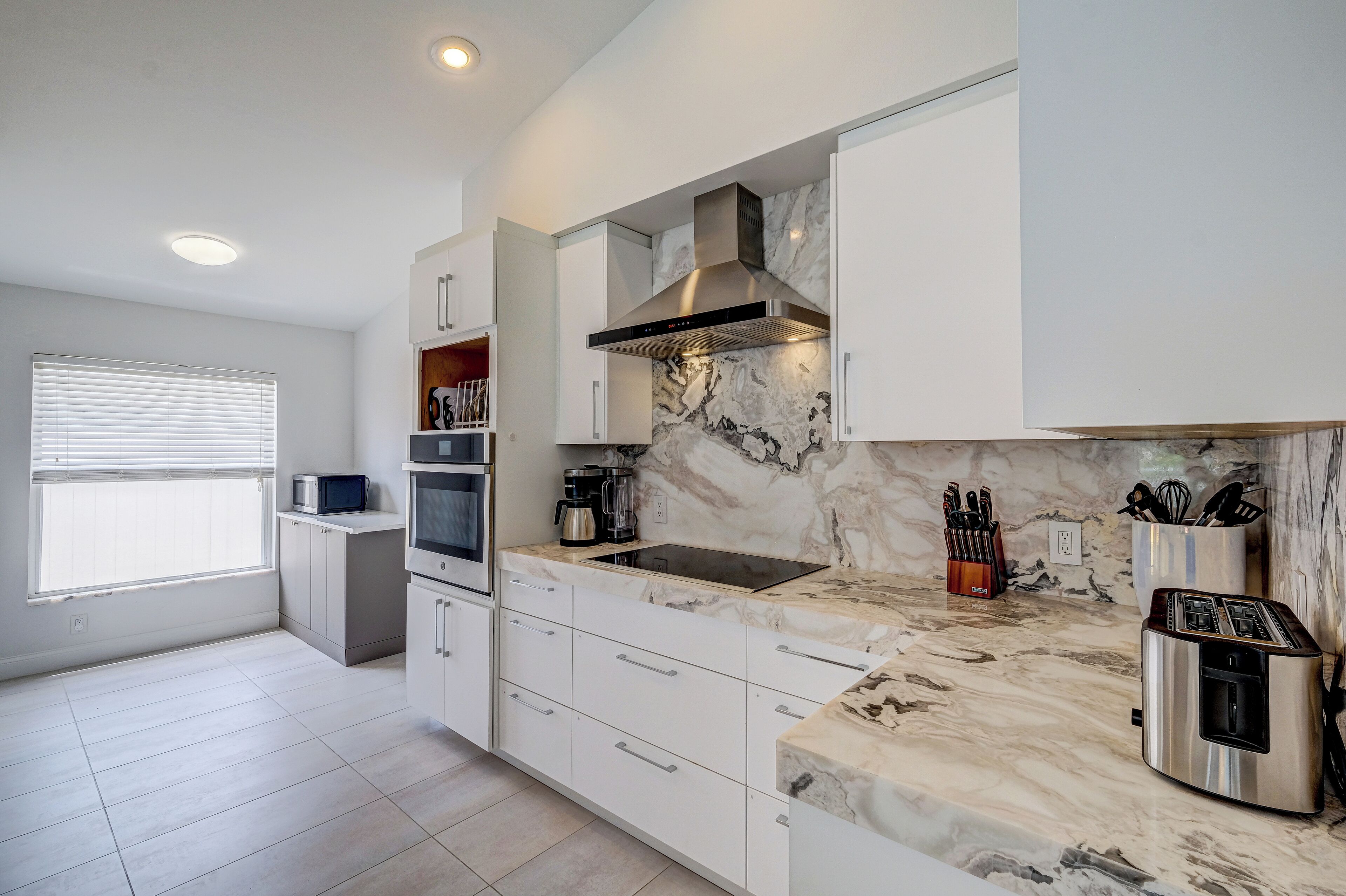 A modern kitchen with white cabinetry, a marble countertop, a stainless steel range hood, and a built-in microwave.