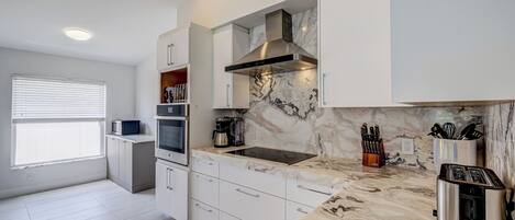 A modern kitchen with white cabinetry, a marble countertop, a stainless steel range hood, and a built-in microwave.