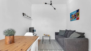 Ein modernes Wohnzimmer mit einem grauen Sofa, einem Holzespeisetisch, einem kleinen Beistelltisch mit Getränken, einer Topfpflanze und einem farbenfrohen, abstrakten Gemälde an der Wand.