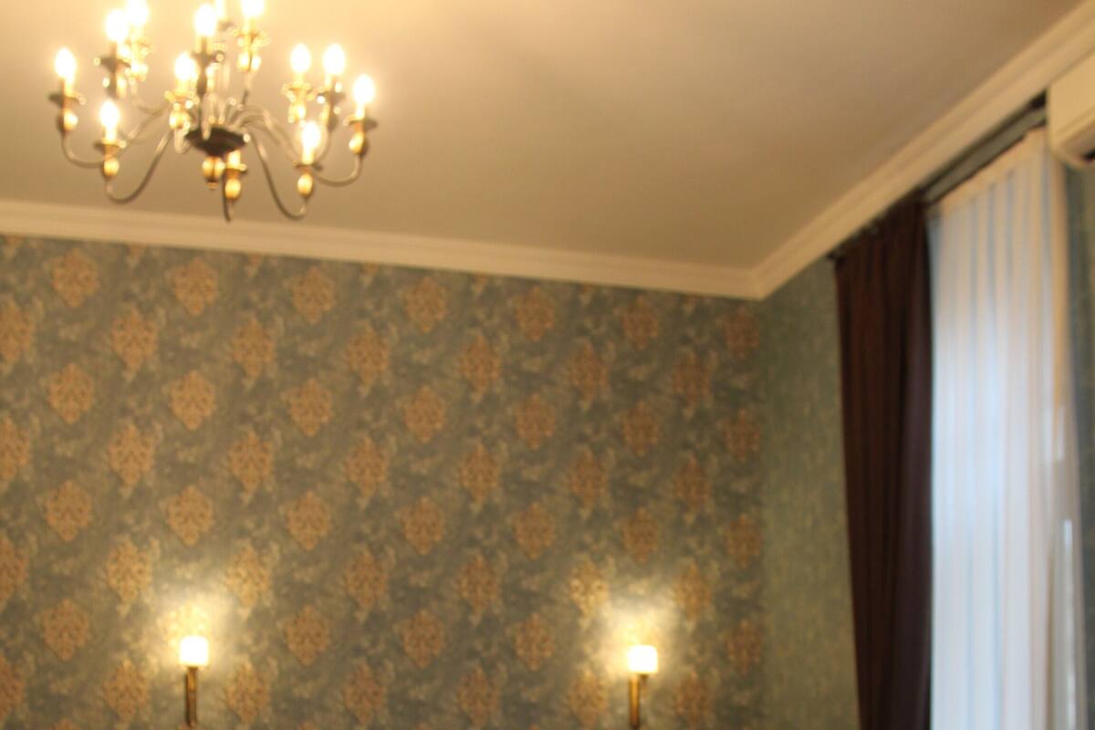 A bedroom with a large bed, a chandelier, patterned wallpaper, and a view of the outdoors through a window.