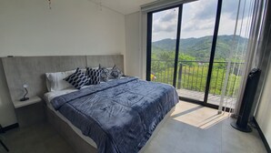 A bedroom with a large bed, a nightstand, a lamp, and a view of a green landscape through a balcony. - Vista Hermosa Glamping & Cabañas (La Mesa)