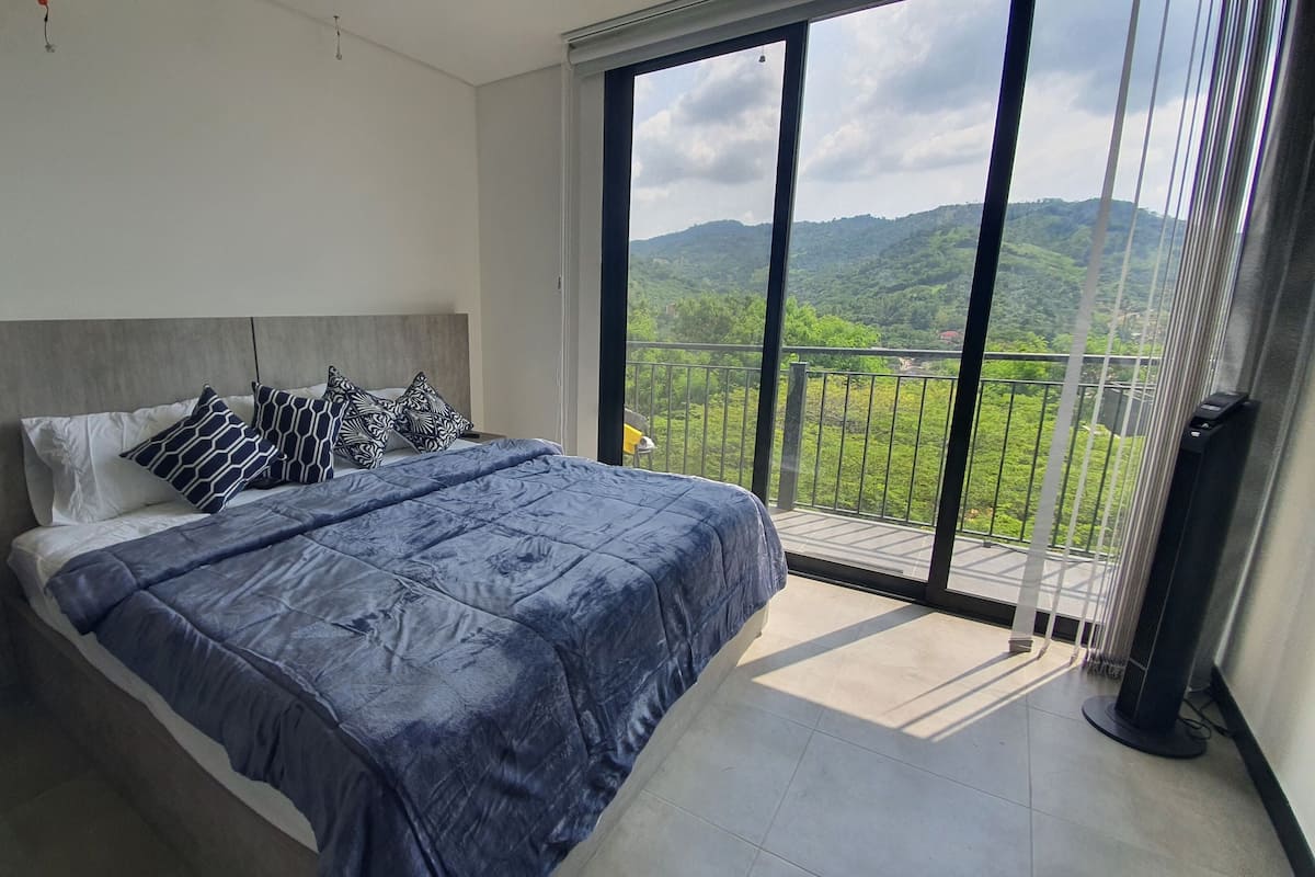 A bedroom with a large bed, a nightstand, a lamp, and a view of a green landscape through a balcony.