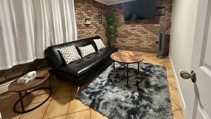 A living room with a black leather sofa, a round wooden coffee table, a fluffy rug, a brick wall, and a mounted TV. - Short Walk to the Falls,SuperLargeApt,Free parking (Niagara Falls)