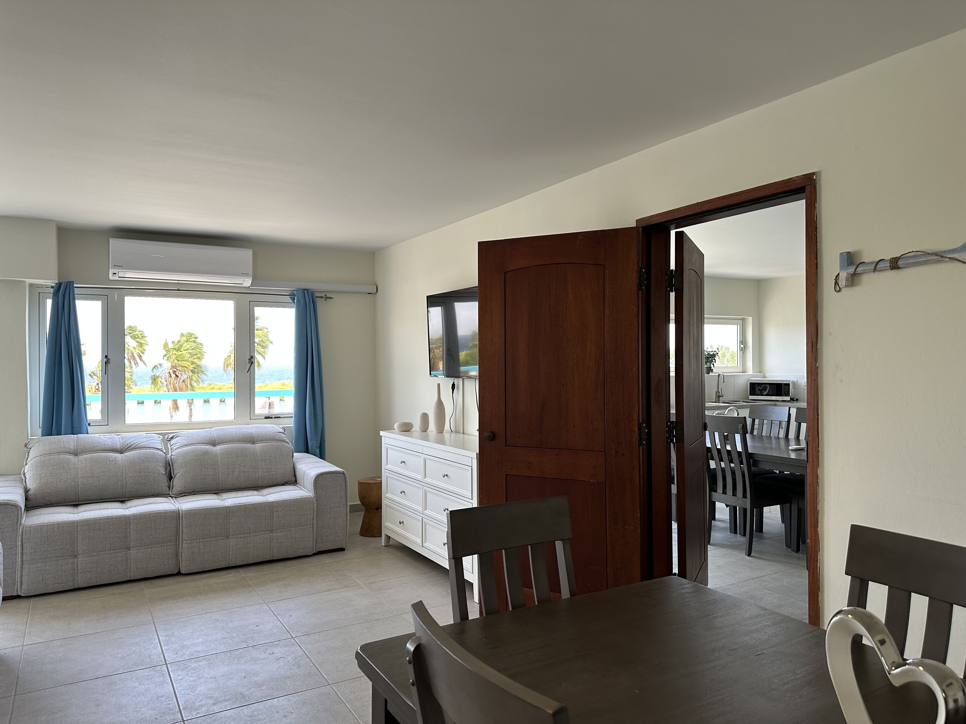 Una sala de estar con un sofá, una mesa de comedor con sillas, una puerta de madera y vistas al océano a través de una ventana.