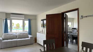 Una sala de estar con un sofá, una mesa de comedor con sillas, una puerta de madera y vistas al océano a través de una ventana.