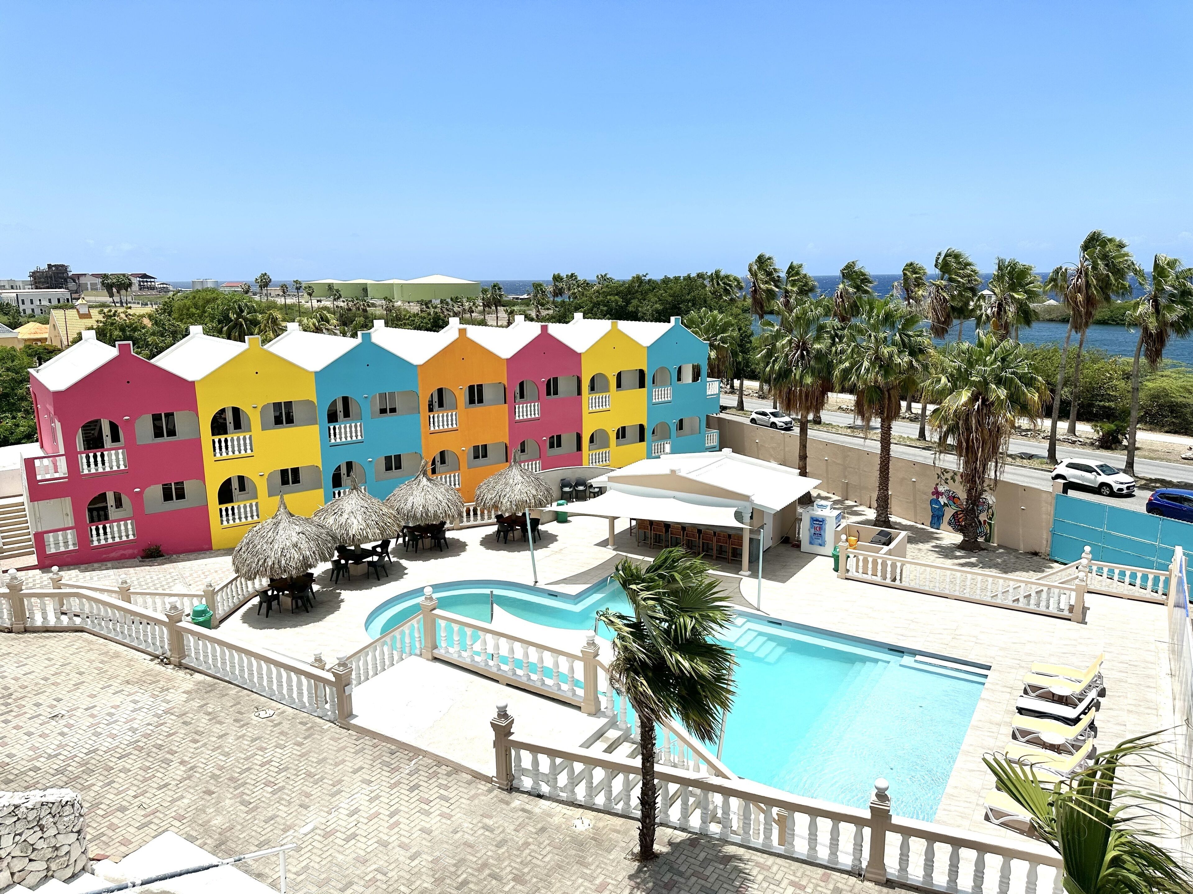 Um hotel colorido com piscina, palmeiras e vista para o mar.