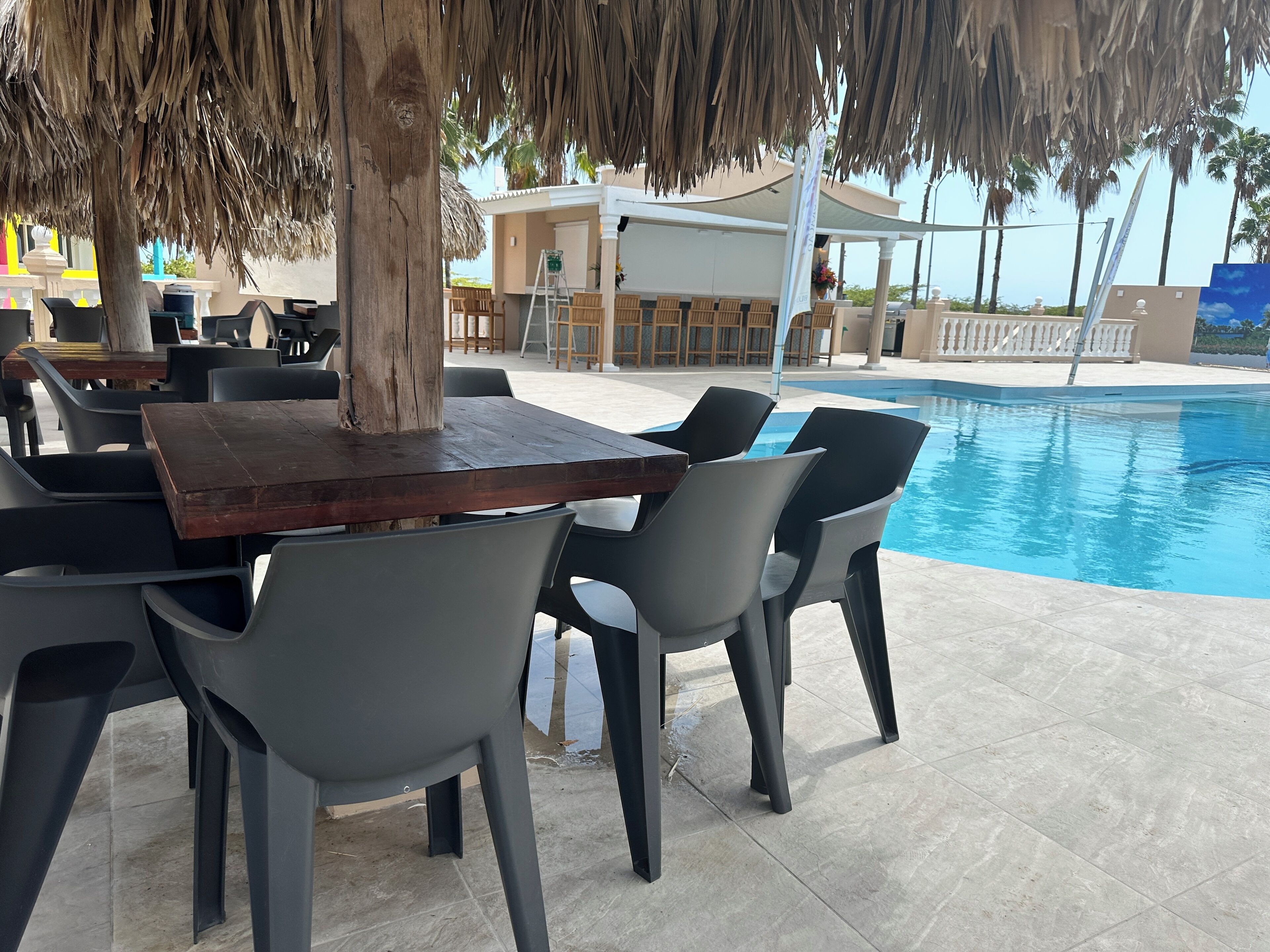 An outdoor seating area with a pool, featuring tables and chairs, a thatched roof, and a view of the pool.