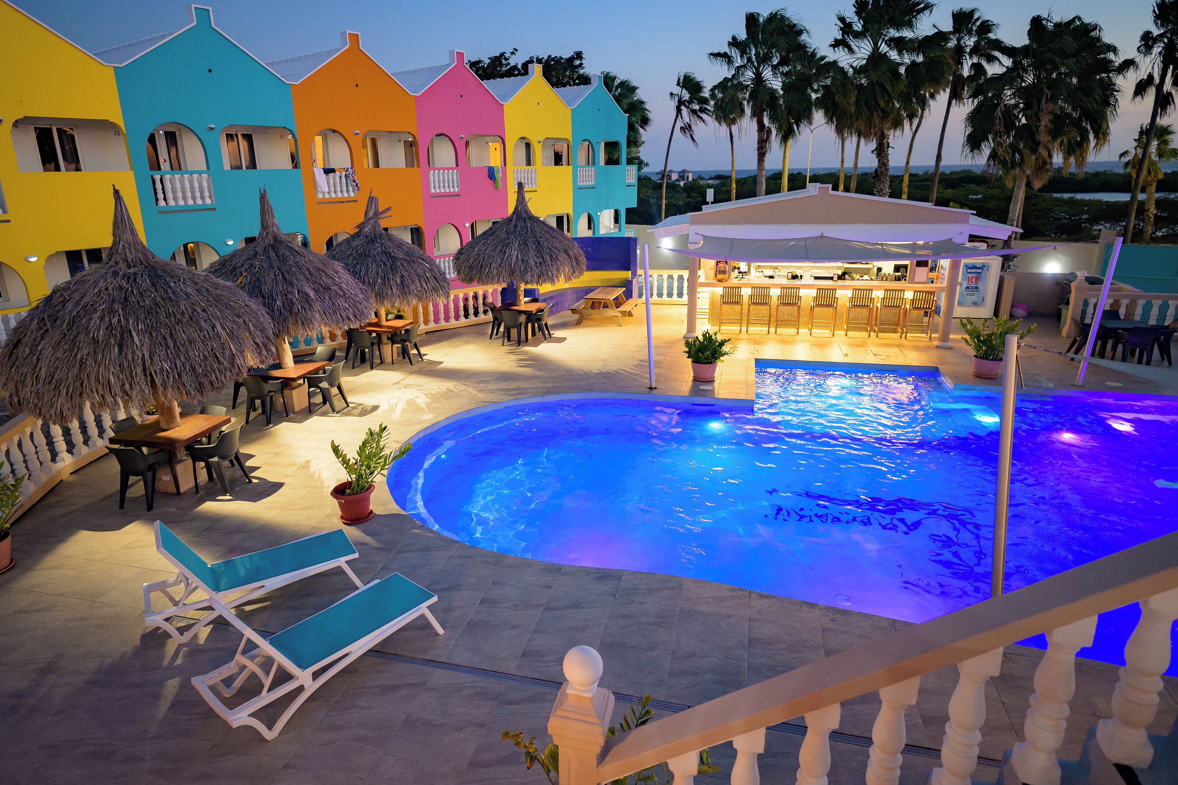 Un área de piscina con sillones de descanso y sombrillas, rodeada de edificios coloridos con balcones.