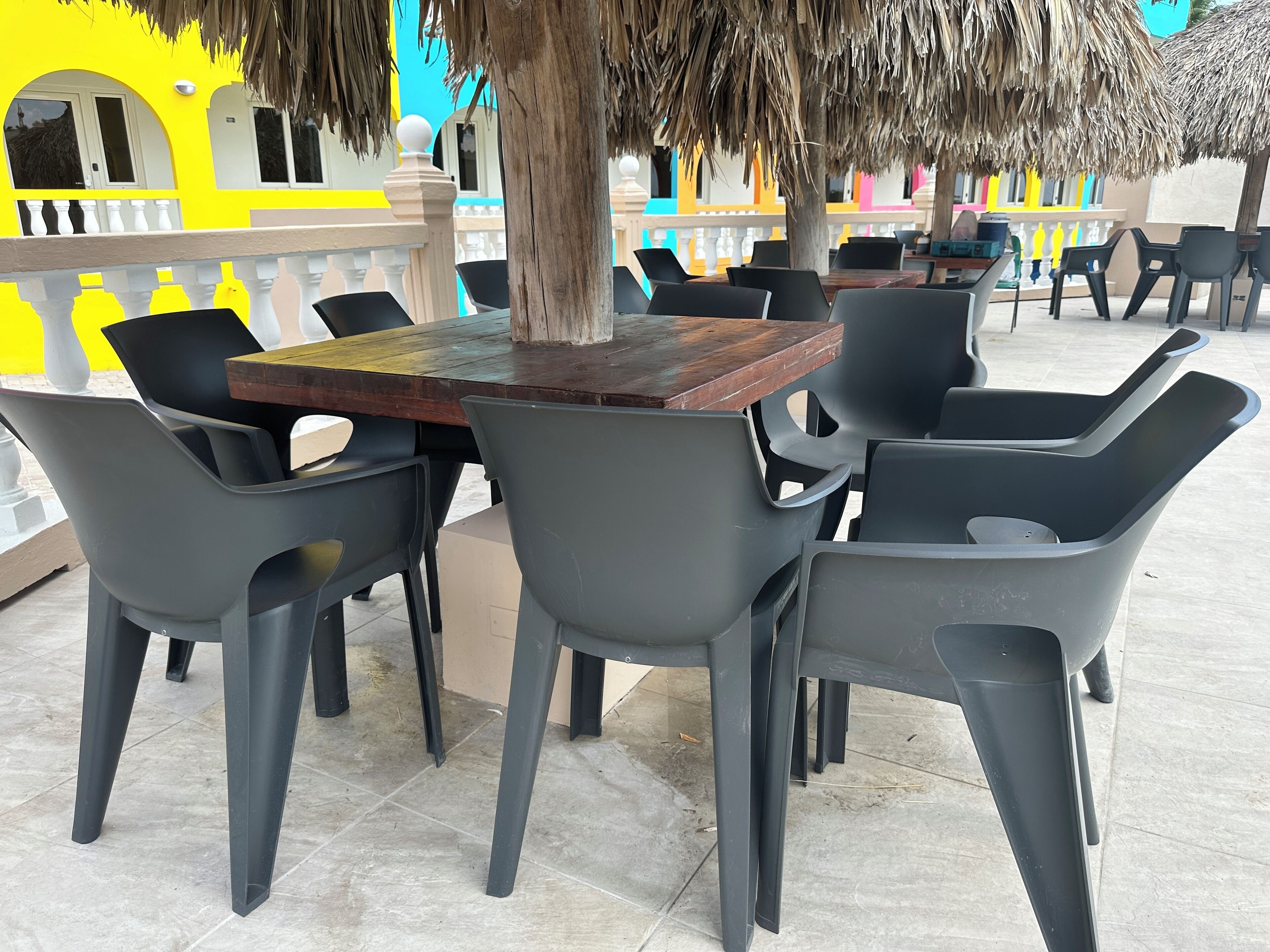 An outdoor seating area with a wooden table and grey plastic chairs, surrounded by a tiled floor and a decorative fence.