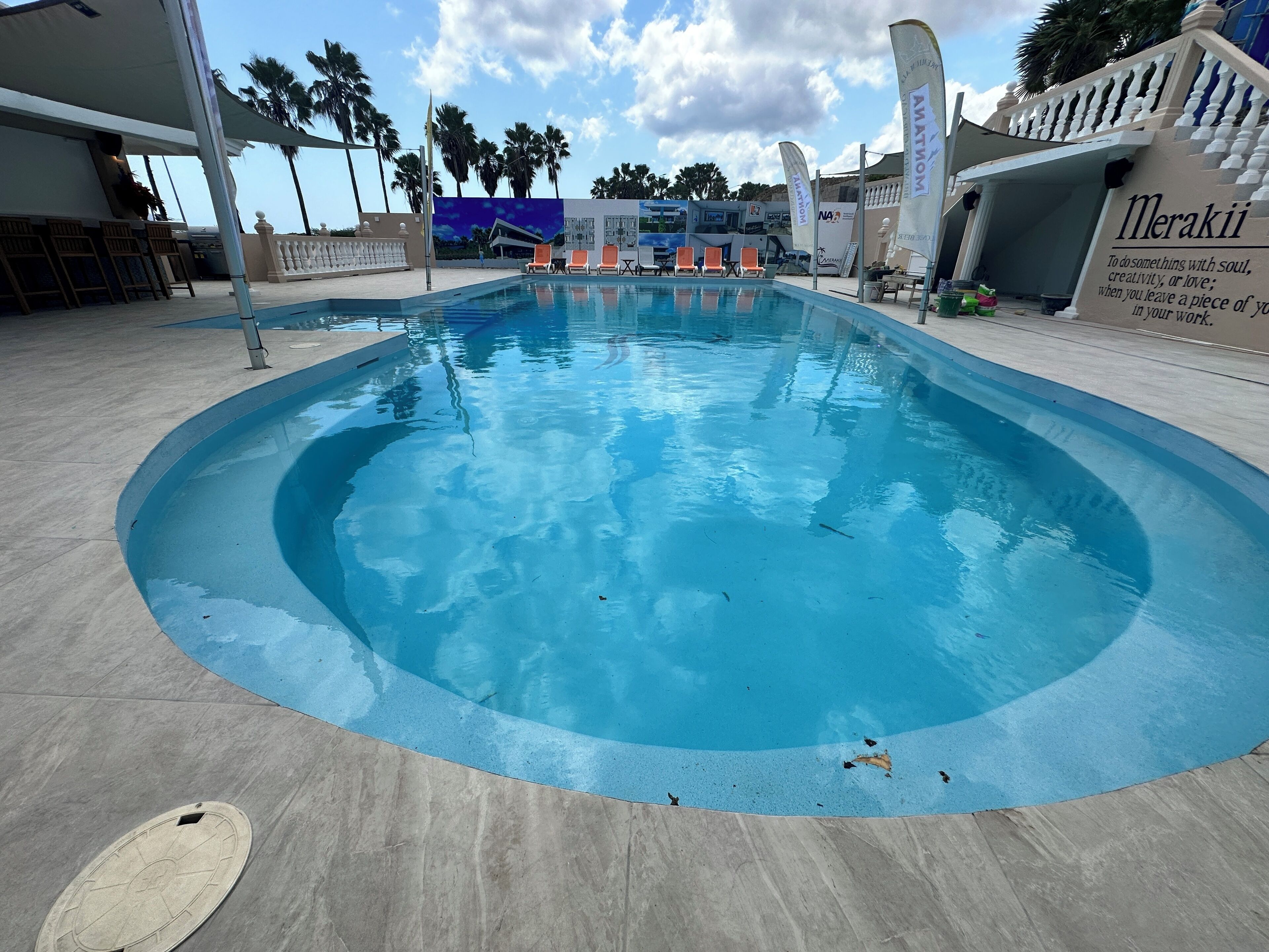 Ein Pool mit gewölbter Kante, umgeben von einer Betonplattform. Im Hintergrund sind Liegestühle und Sonnenschirme zu sehen.