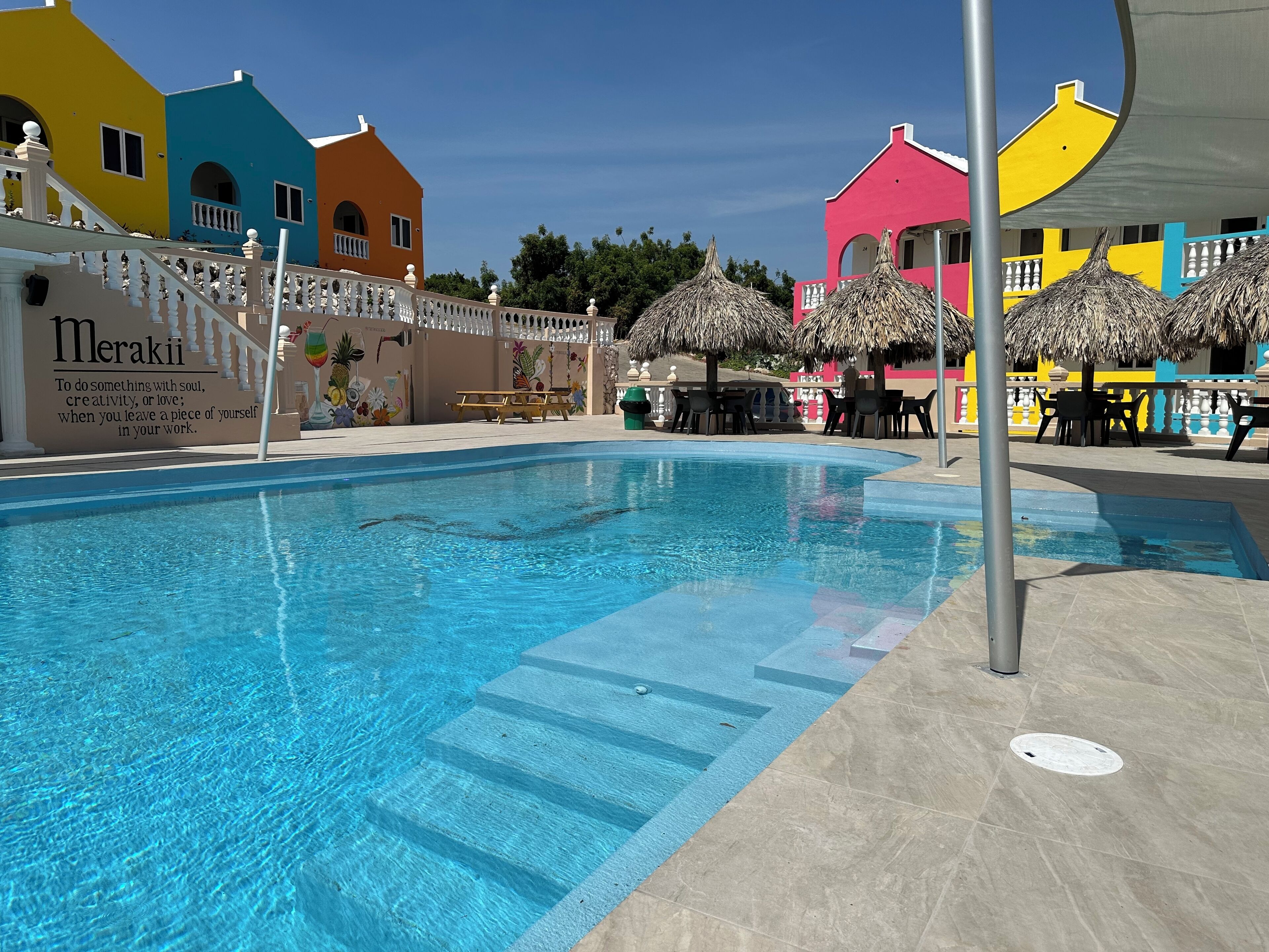 Une piscine entourée d’une clôture blanche, avec des bâtiments aux couleurs vives en arrière-plan.