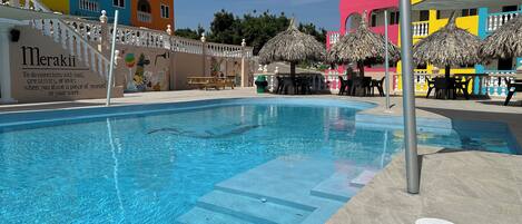 Une piscine entourée d’une clôture blanche, avec des bâtiments aux couleurs vives en arrière-plan.