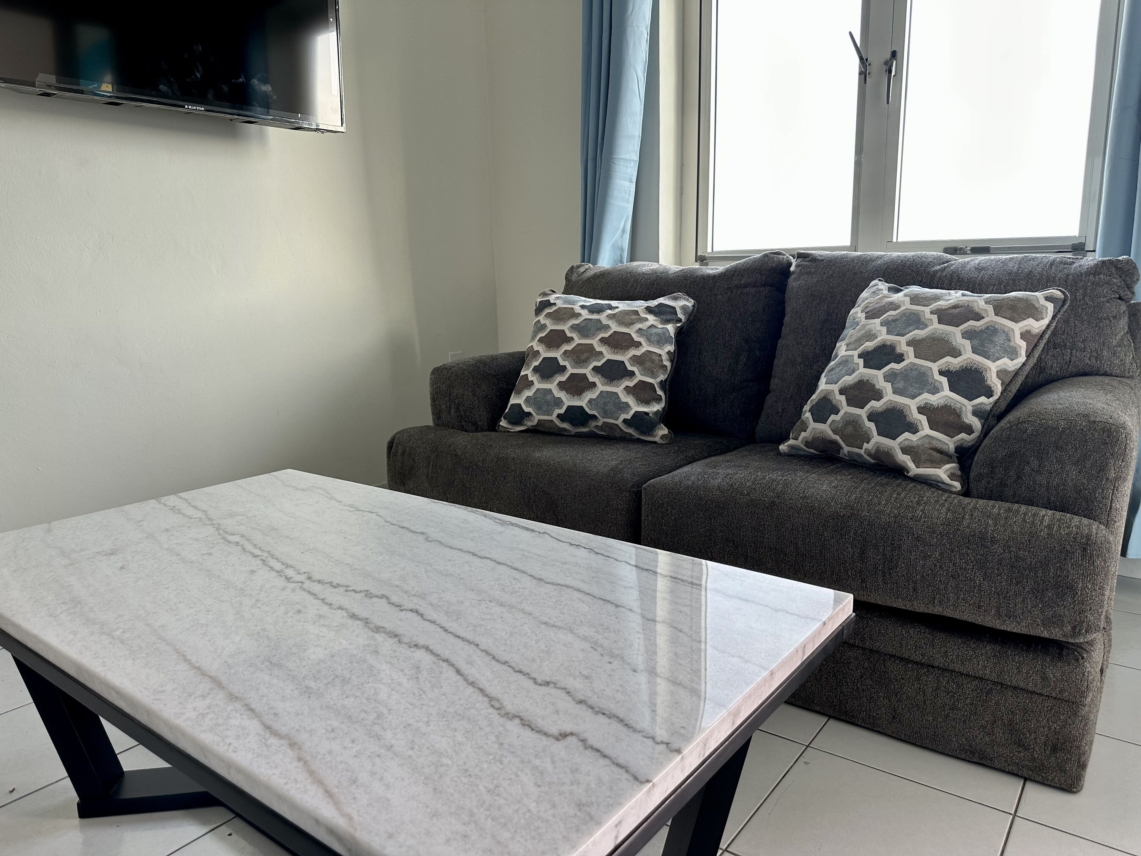 Uma sala de estar com um sofá cinza, almofadas estampadas, uma mesa de centro de mármore e uma TV de tela plana fixada na parede.
