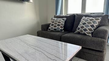 Uma sala de estar com um sofá cinza, almofadas estampadas, uma mesa de centro de mármore e uma TV de tela plana fixada na parede.